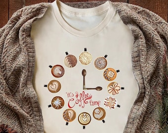 May include: Cream-colored t-shirt featuring a coffee-themed design. The design includes coffee cup illustrations arranged in a clock formation, with a spoon forming the clock hands. The text "It's Coffee Time" is written in red script.