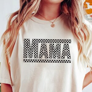 Puede incluir: Camiseta color crema con la palabra "MAMA" en un dise&ntilde;o a cuadros blanco y negro. La camiseta es de corte holgado y tiene mangas cortas. La modelo lleva un pantal&oacute;n corto verde oscuro y una pulsera.