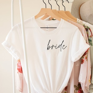 May include: White t-shirt with the word "bride" written in black script on the front. The shirt is hanging on a white rack with other clothing.