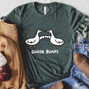 May include: A green t-shirt with a white goose graphic and the text "Goose Bumps". The shirt is displayed on a white surface with a brown sweater, blue jeans, and brown boots.