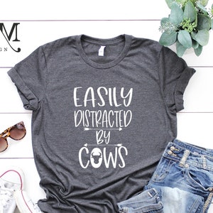 May include: Dark gray t-shirt with the text "EASILY DISTRACTED BY COWS" in white. The shirt is paired with blue jeans, white sneakers, and sunglasses. A sprig of greenery adds a decorative touch.