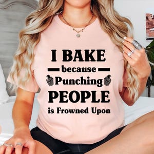 May include: A heather peach t-shirt featuring black text that reads I BAKE because Punching PEOPLE is Frowned Upon, with small cupcake graphics on either side of the word Punching. The shirt has a crew neckline and short sleeves.