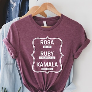 May include: A maroon t-shirt with a white graphic design that reads "ROSA SAT. SO RUBY COULD WALK. SO KAMALA COULD RUN."