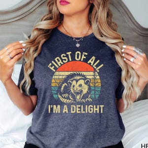 May include: A dark heather navy blue t-shirt with a vintage-style graphic of a possum with the text "First of all I'm a delight".