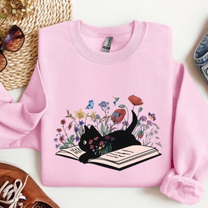 May include: Pink sweatshirt with a black cat reading a book surrounded by flowers and butterflies. The cat is sitting on an open book with pages.