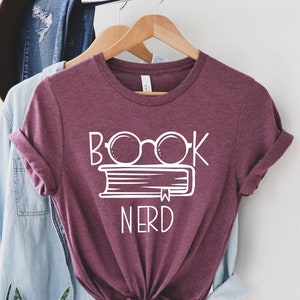 May include: A maroon t-shirt with the words "BOOK NERD" in white, featuring a pair of glasses over a book. The shirt is hanging on a wooden hanger, with a light blue shirt and a brown hat in the background.