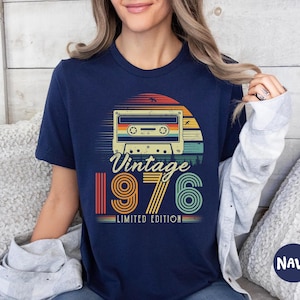 May include: A navy blue short-sleeve t-shirt featuring a graphic of a cassette tape centred above the text "Vintage 1976 LIMITED EDITION". The design incorporates a sunset motif with horizontal stripes in shades of orange, yellow, and teal.