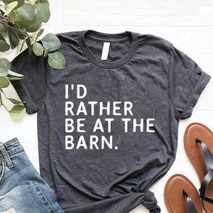 Puede incluir: Camiseta gris oscuro con el texto "I'D RATHER BE AT THE BARN." La camiseta se combina con vaqueros azules y sandalias marrones. Ramas de hojas verdes est&aacute;n dispuestas en la parte superior izquierda de la imagen.