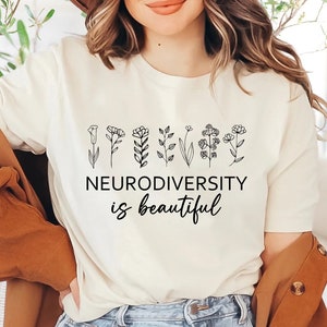 May include: Cream-colored t-shirt with the text "NEURODIVERSITY is beautiful" in black script. Above the text is a line of delicate, black line-drawn flowers. The shirt is being worn with a brown jacket.