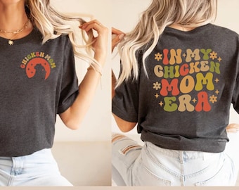 Könnte beinhalten: Ein dunkelgraues T-Shirt mit einem Aufdruck auf der Vorderseite, der "Chicken Mom" in einem Regenbogen-Farbschema zeigt. Die Rückseite des Shirts hat einen Aufdruck, der "In My Chicken Mom Era" in einem Regenbogen-Farbschema zeigt.