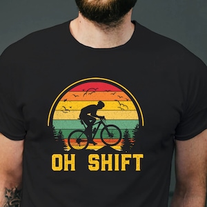 May include: Black t-shirt featuring a cyclist silhouette riding a bicycle against a sunset backdrop. The design includes a mountain scene, birds, and the text "OH SHIFT" in bold, yellow letters. The shirt is made of a soft material.
