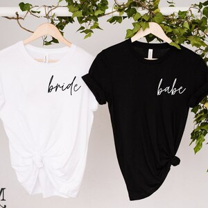 K&ouml;nnte beinhalten: Zwei T-Shirts auf Holzb&uuml;geln. Eines ist wei&szlig; mit dem Wort "bride" in schwarzer Schrift. Das andere ist schwarz mit dem Wort "babe" in wei&szlig;er Schrift. Gr&uuml;nes Laub befindet sich im Hintergrund.