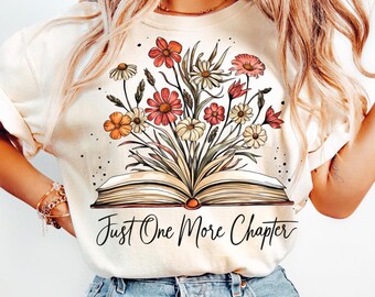 May include: A cream-colored t-shirt featuring a graphic of an open book with a bouquet of colorful flowers emerging from the pages. Below the book, the text reads Just One More Chapter in a black cursive script. The shirt has short sleeves.