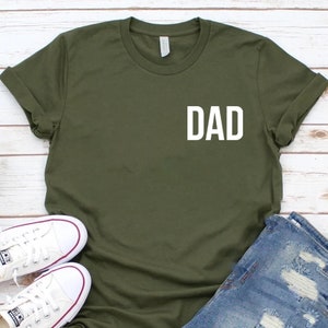 May include: A green t-shirt with the word "DAD" printed in white on the front. The shirt is laid flat on a white wooden surface with a pair of white sneakers and a pair of blue denim shorts.