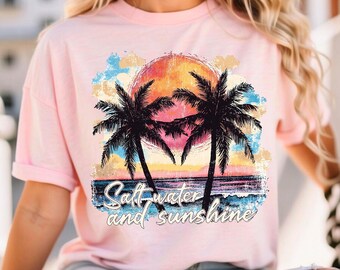 May include: A light pink t-shirt featuring a tropical sunset design with palm trees and the text "Saltwater and Sunshine". The graphic includes a vibrant orange and yellow sun, blue ocean waves, and colorful clouds.