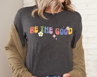 May include: A dark charcoal gray short-sleeve t-shirt featuring the text BE THE GOOD in colorful, wavy, retro-style lettering. The design includes two white daisy flowers with yellow centers and two small pink four-pointed star icons.