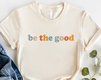 May include: Cream-colored t-shirt with the phrase "be the good" in colorful, retro-style lettering. The text is in shades of blue, orange, and brown. The shirt has short sleeves and a crew neck.