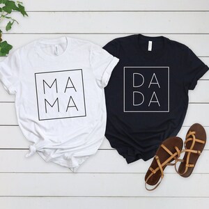 May include: Two white t-shirts with black text. One shirt says "MA MA" in a square and the other says "DA DA" in a square.