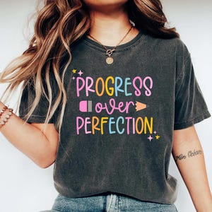 May include: Dark gray t-shirt with the words "PROGRESS over PERFECTION" in colorful, playful lettering. The design includes a pencil and a star graphic. The shirt is a comfortable, casual style.