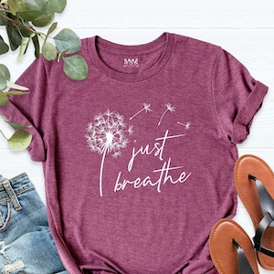 May include: A maroon t-shirt with a white dandelion graphic and the words "just breathe." The shirt is paired with blue jeans and brown sandals with black straps. The t-shirt is folded at the bottom.