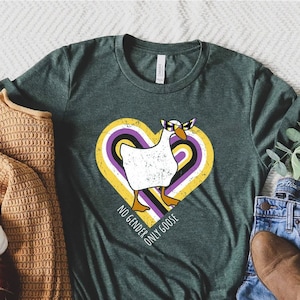 May include: A green t-shirt with a white goose wearing sunglasses in a heart with purple, yellow, and black stripes. The text on the shirt reads "No Gender, Only Goose".