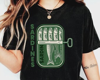 Puede incluir: Una camiseta negra con un gráfico verde de una lata de sardinas abierta. El diseño incluye el texto SARDINES en vertical, y SARDIN, IN PURE OUVE y SARDINE FILLET IS INC dentro de la ilustración de la lata, que muestra cinco peces estilizados.