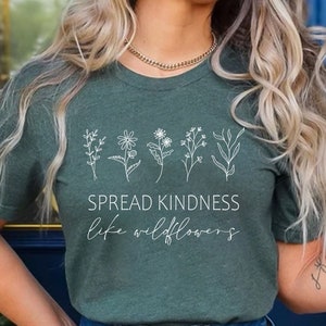 Op de afbeelding: Een groen T-shirt met een witte afbeelding van wilde bloemen en de tekst "Spread Kindness Like Wildflowers".