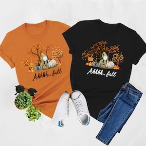 May include: An orange t-shirt and a black t-shirt, each featuring a graphic of a gnome, pumpkins, a black cat, and autumn trees with the text Ahhhh...fall. The shirts are displayed with black sunglasses, a small green plant, and blue jeans.