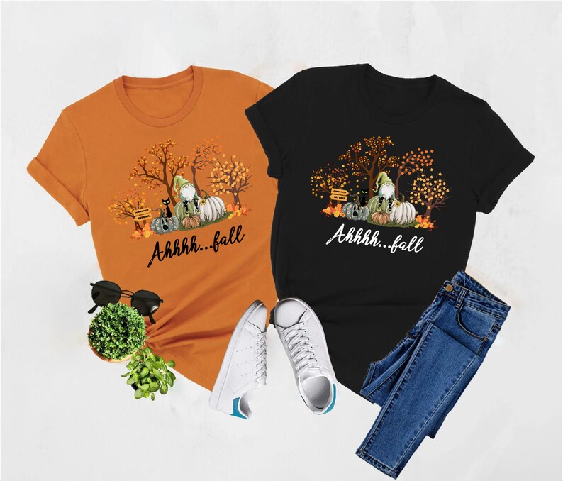 May include: An orange t-shirt and a black t-shirt, each featuring a graphic of a gnome, pumpkins, a black cat, and autumn trees with the text Ahhhh...fall. The shirts are displayed with black sunglasses, a small green plant, and blue jeans.