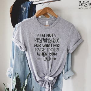 以下が含まれることがあります： クルーネックと結び目のある裾が特徴のヘザーグレーの半袖Tシャツ。前面には「I'M NOT RESPONSIBLE FOR WHAT MY FACE DOES WHEN YOU TALK」という黒いテキストがプリントされており、小さな矢印の装飾が施されています。