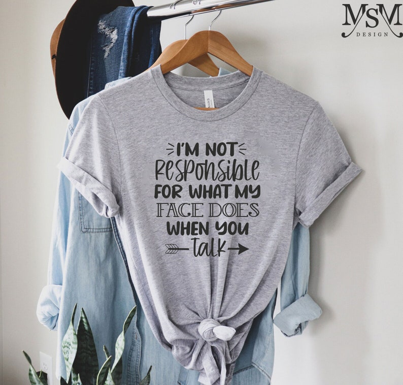 以下が含まれることがあります： クルーネックと結び目のある裾が特徴のヘザーグレーの半袖Tシャツ。前面には「I'M NOT RESPONSIBLE FOR WHAT MY FACE DOES WHEN YOU TALK」という黒いテキストがプリントされており、小さな矢印の装飾が施されています。