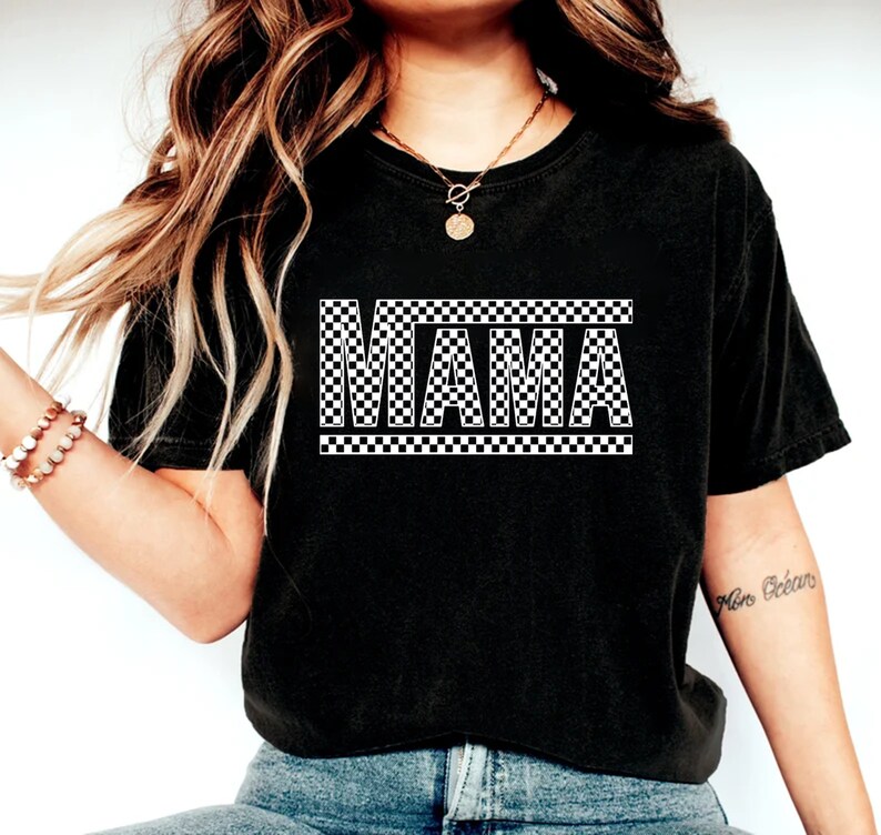 Puede incluir: Camiseta negra con la palabra "MAMA" en un patr&oacute;n de cuadros blancos y negros. La camiseta es de estilo casual, adecuada para el uso diario. El dise&ntilde;o est&aacute; centrado en la parte delantera.