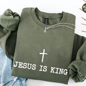 K&ouml;nnte beinhalten: Ein olivgr&uuml;nes Sweatshirt mit dem Text "JESUS IS KING" und einem wei&szlig;en Kreuz. Eine silberne Halskette mit einem kreisf&ouml;rmigen Anh&auml;nger liegt auf dem Sweatshirt. Das Kleidungsst&uuml;ck ist gefaltet, mit den &Auml;rmeln und einer blauen Jeans sichtbar.