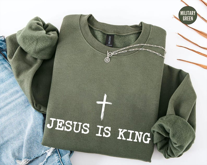 K&ouml;nnte beinhalten: Ein olivgr&uuml;nes Sweatshirt mit dem Text "JESUS IS KING" und einem wei&szlig;en Kreuz. Eine silberne Halskette mit einem kreisf&ouml;rmigen Anh&auml;nger liegt auf dem Sweatshirt. Das Kleidungsst&uuml;ck ist gefaltet, mit den &Auml;rmeln und einer blauen Jeans sichtbar.