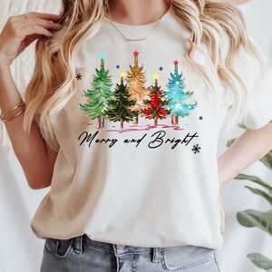Pu&ograve; includere: Una maglietta bianca con un design di albero di Natale e il testo "Merry and Bright". Gli alberi sono decorati con luci colorate e ornamenti.