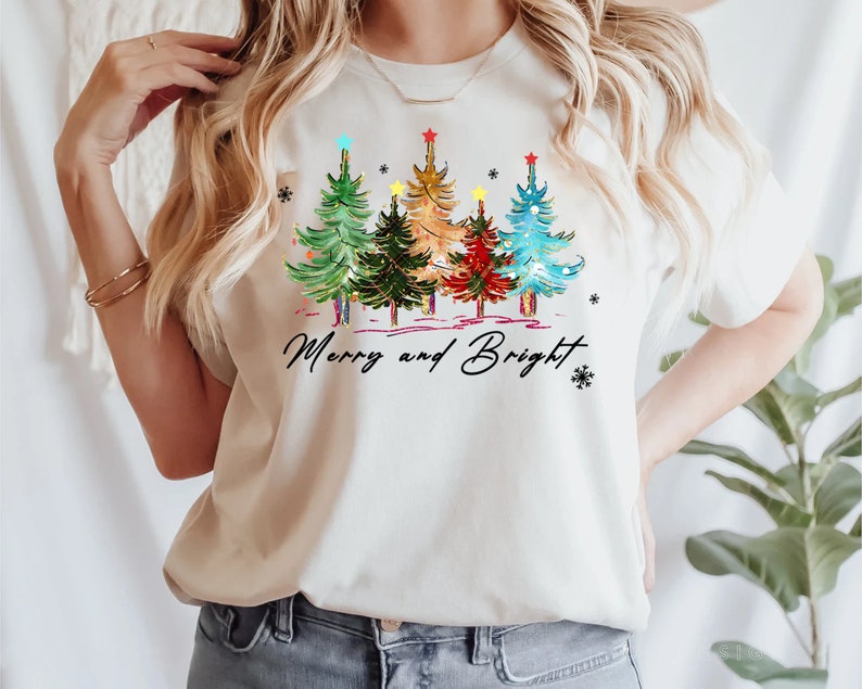 Pu&ograve; includere: Una maglietta bianca con un design di albero di Natale e il testo "Merry and Bright". Gli alberi sono decorati con luci colorate e ornamenti.