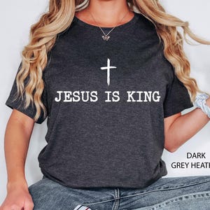 K&ouml;nnte beinhalten: Dunkelgraues meliertes T-Shirt mit dem wei&szlig;en Text &bdquo;JESUS IS KING&ldquo; und einem wei&szlig;en Kreuz dar&uuml;ber. Das T-Shirt hat einen Rundhalsausschnitt und kurze &Auml;rmel.