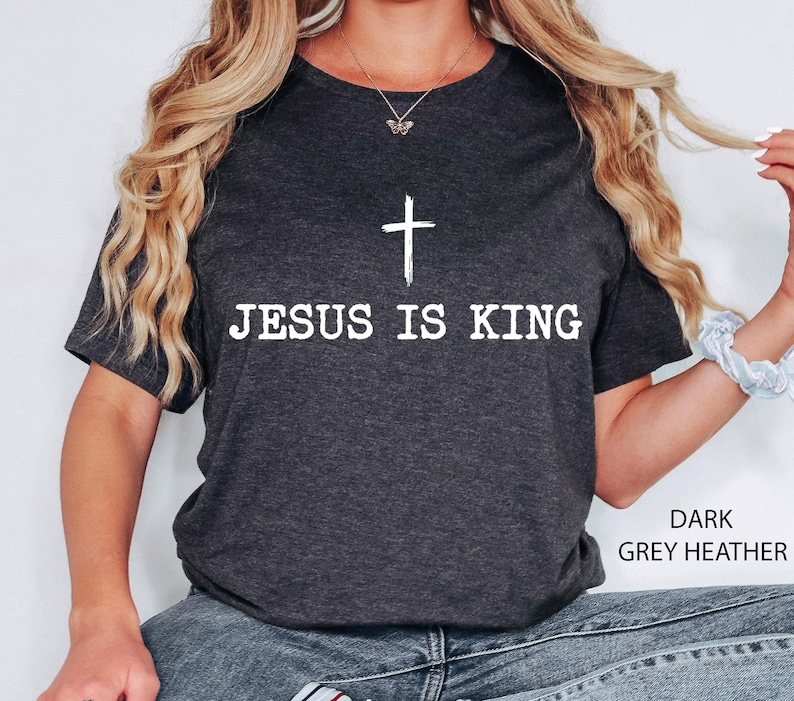K&ouml;nnte beinhalten: Dunkelgraues meliertes T-Shirt mit dem wei&szlig;en Text &bdquo;JESUS IS KING&ldquo; und einem wei&szlig;en Kreuz dar&uuml;ber. Das T-Shirt hat einen Rundhalsausschnitt und kurze &Auml;rmel.