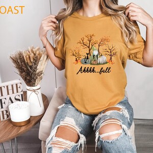 May include: A mustard yellow short-sleeved t-shirt featuring a graphic print of autumn trees, pumpkins, a black cat, and a gnome. The text "Ahhhh...fall" is printed in black script below the graphic. The shirt is paired with distressed blue jeans.