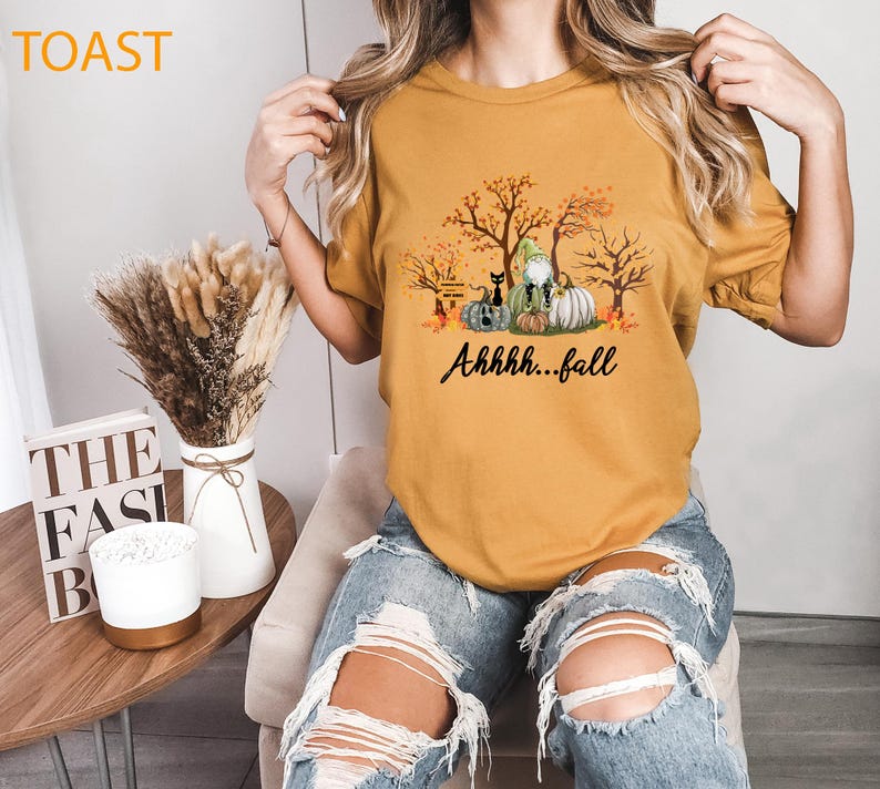 May include: A mustard yellow short-sleeved t-shirt featuring a graphic print of autumn trees, pumpkins, a black cat, and a gnome. The text "Ahhhh...fall" is printed in black script below the graphic. The shirt is paired with distressed blue jeans.