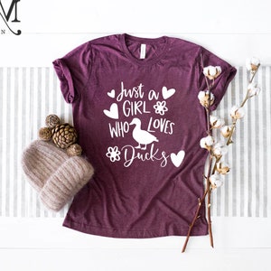 May include: A maroon t-shirt with the text "Just a girl who loves ducks" in white with a duck illustration and hearts.