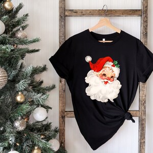 May include: Black t-shirt featuring a vintage Santa Claus graphic. The Santa Claus illustration has a red hat, white beard, and rosy cheeks. The shirt is hanging on a wooden hanger against a white background, with a Christmas tree on the left.