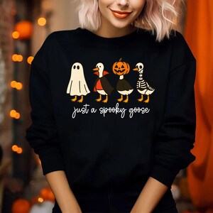 Puede incluir: Sudadera negra con un dise&ntilde;o de Halloween. El gr&aacute;fico muestra cuatro gansos disfrazados de fantasma, vampiro, calabaza y esqueleto. La frase "just a spooky goose" est&aacute; escrita en cursiva debajo.