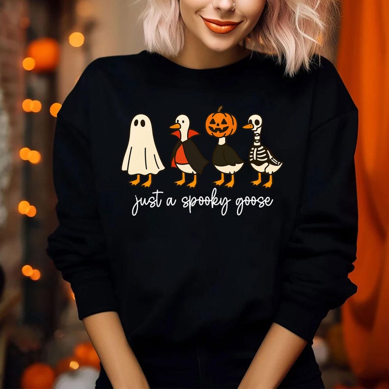 Puede incluir: Sudadera negra con un dise&ntilde;o de Halloween. El gr&aacute;fico muestra cuatro gansos disfrazados de fantasma, vampiro, calabaza y esqueleto. La frase "just a spooky goose" est&aacute; escrita en cursiva debajo.
