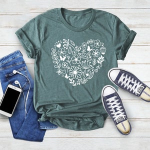 May include: A teal green t-shirt with a white floral heart design. The shirt is laid flat on a white wooden surface with a pair of blue jeans, a phone, and headphones. There are also a pair of dark blue sneakers with white laces and the word "Converse" on the side.