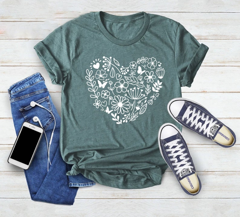 May include: A teal green t-shirt with a white floral heart design. The shirt is laid flat on a white wooden surface with a pair of blue jeans, a phone, and headphones. There are also a pair of dark blue sneakers with white laces and the word "Converse" on the side.