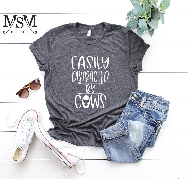 May include: Dark gray t-shirt with the text "EASILY DISTRACTED BY COWS" in white. The shirt is paired with blue jeans, white sneakers, and sunglasses. A sprig of greenery adds a decorative touch.