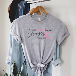 May include: A heather gray t-shirt with the text "The Stronger is Always Than the Struggle" in black and pink. The shirt is hanging on a wooden hanger.