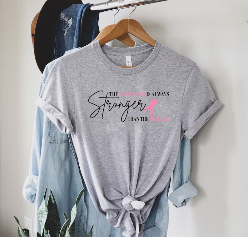 May include: A heather gray t-shirt with the text "The Stronger is Always Than the Struggle" in black and pink. The shirt is hanging on a wooden hanger.