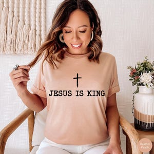 K&ouml;nnte beinhalten: Ein pfirsichfarbenes T-Shirt mit dem Text "JESUS IS KING" in Schwarz aufgedruckt, mit einem einfachen Kreuz &uuml;ber dem Text. Das Shirt hat kurze &Auml;rmel und einen Rundhalsausschnitt. Das Model sitzt auf einem Holzstuhl.